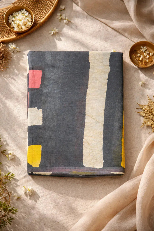 Folded soft silk saree with abstract patterns in black, white, yellow, and pink, styled on beige fabric with floral accents.