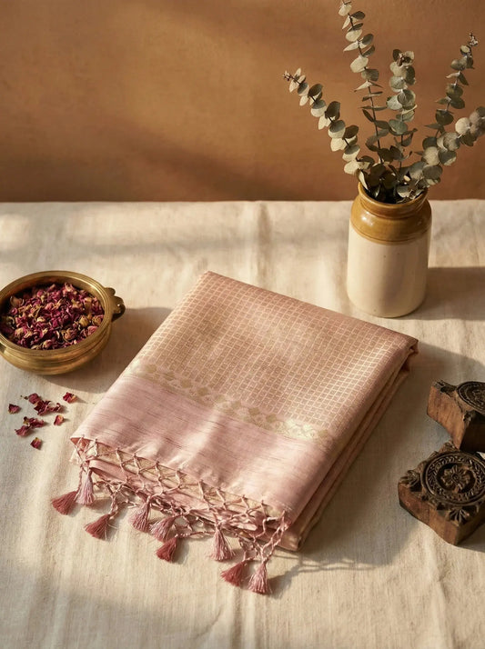 Soft pink silk saree with gold border and tassels, displayed on a beige cloth with dried flowers and a ceramic vase
