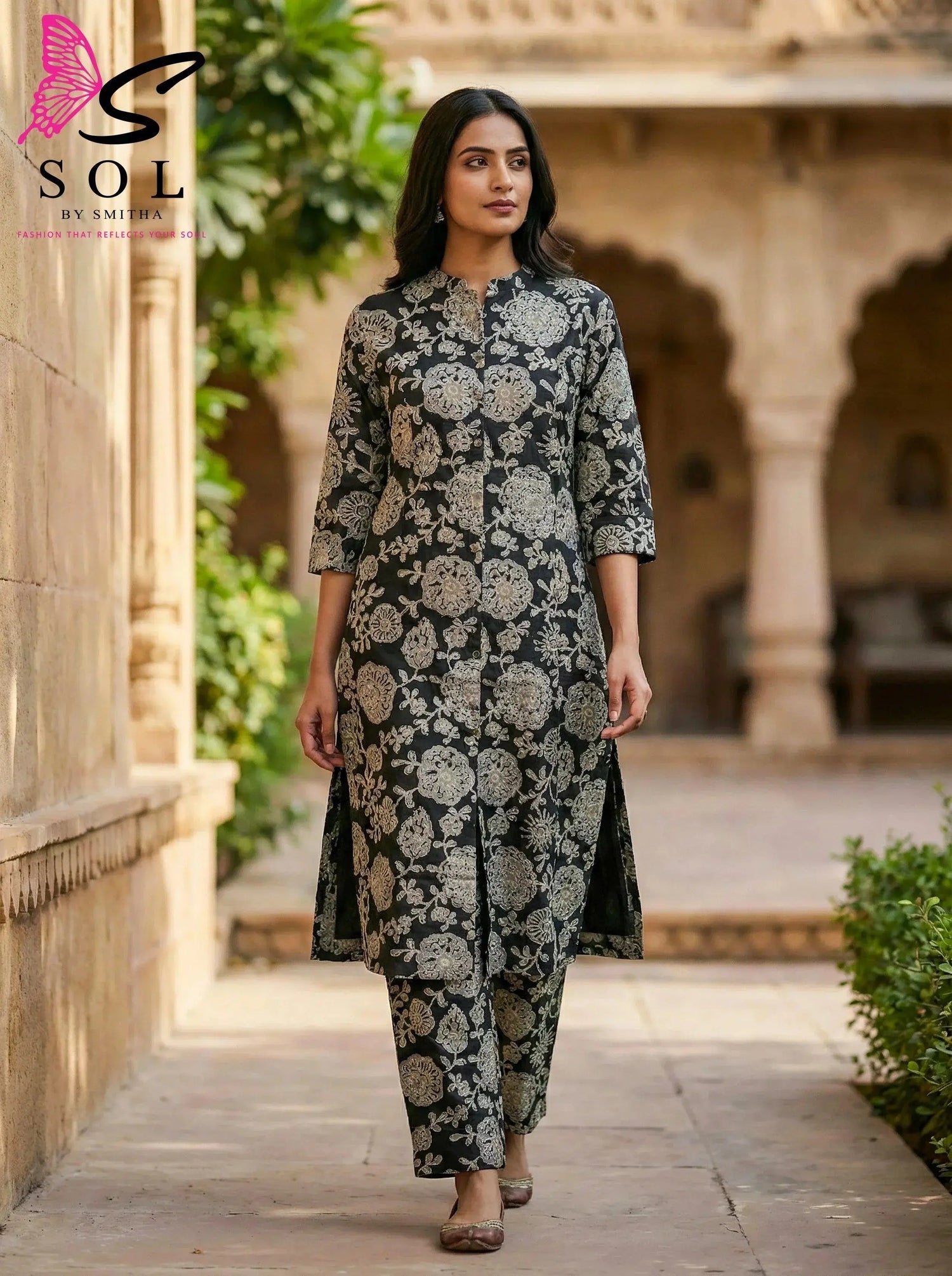 Woman wearing a black and beige floral silk kurta set, walking in a traditional courtyard.