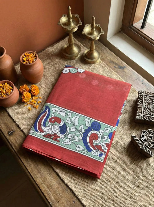 Red soft silk saree with peacock and floral border, displayed on rustic wooden table with brass lamps and clay pots