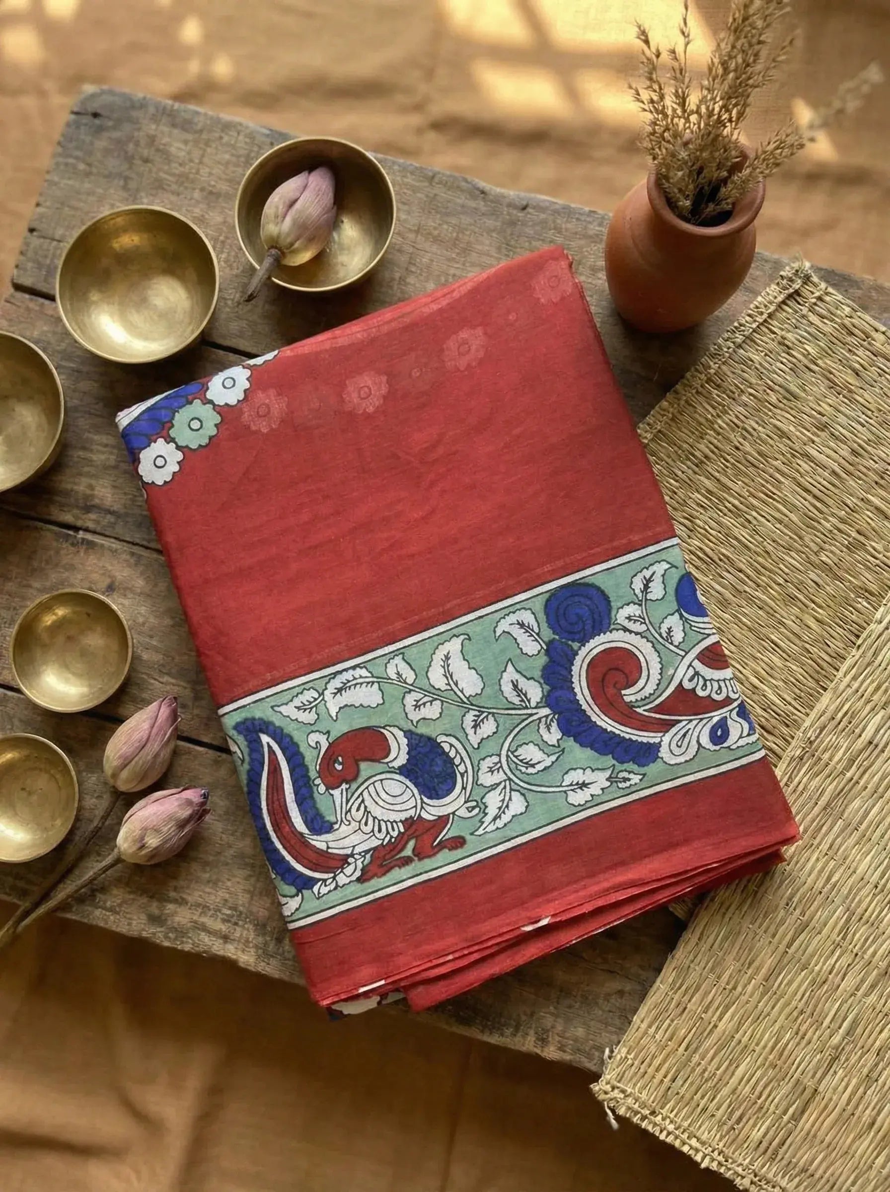 Red soft silk saree with peacock and floral border, styled on rustic wooden table with brass bowls and lotus buds.