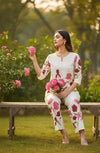Woman in white floral ethnic outfit holding pink flowers, seated on a bench in a garden