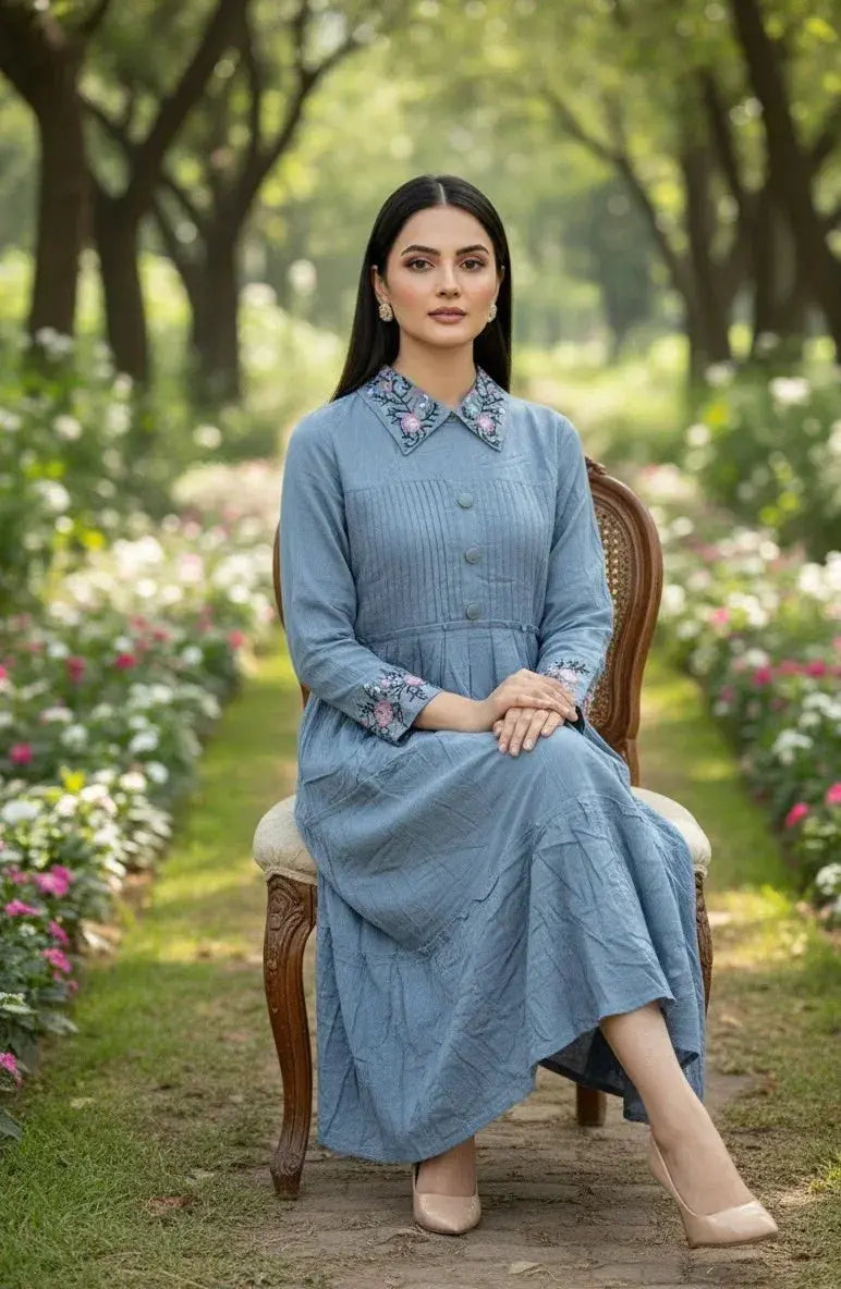 Woman in a blue embroidered long dress with floral cuffs and collar, sitting in a garden.