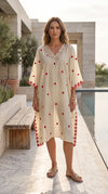 A woman stands outdoors by a pool wearing a loose cream kaftan dress with red embroidered motifs and a beaded neckline. The setting includes a stone patio, bench, and modern building architecture.