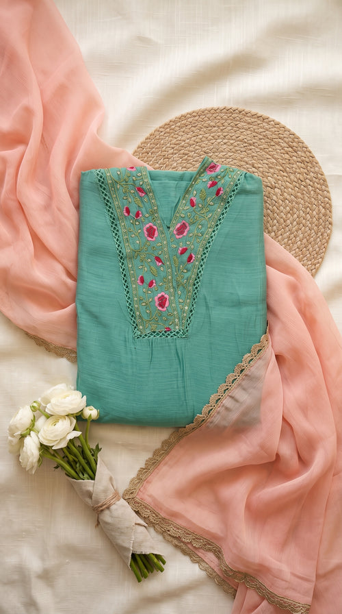 A folded mint green kurta with pink and gold floral embroidery on the neckline, styled with a peach-pink dupatta, a woven straw hat, and white flowers on a cream fabric background.