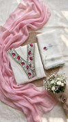 A styled flat-lay of traditional Indian clothing and accessories, featuring a white kurta with pink floral embroidery and green borders, a matching folded white dupatta, a draped pink scarf, and a small bouquet of white baby's breath on a neutral fabric background.