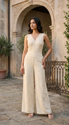A woman models a sleeveless, wide-leg jumpsuit in a warm cream or off-white color with a subtle textured jacquard pattern. She is standing outdoors in front of a stone archway and courtyard setting.