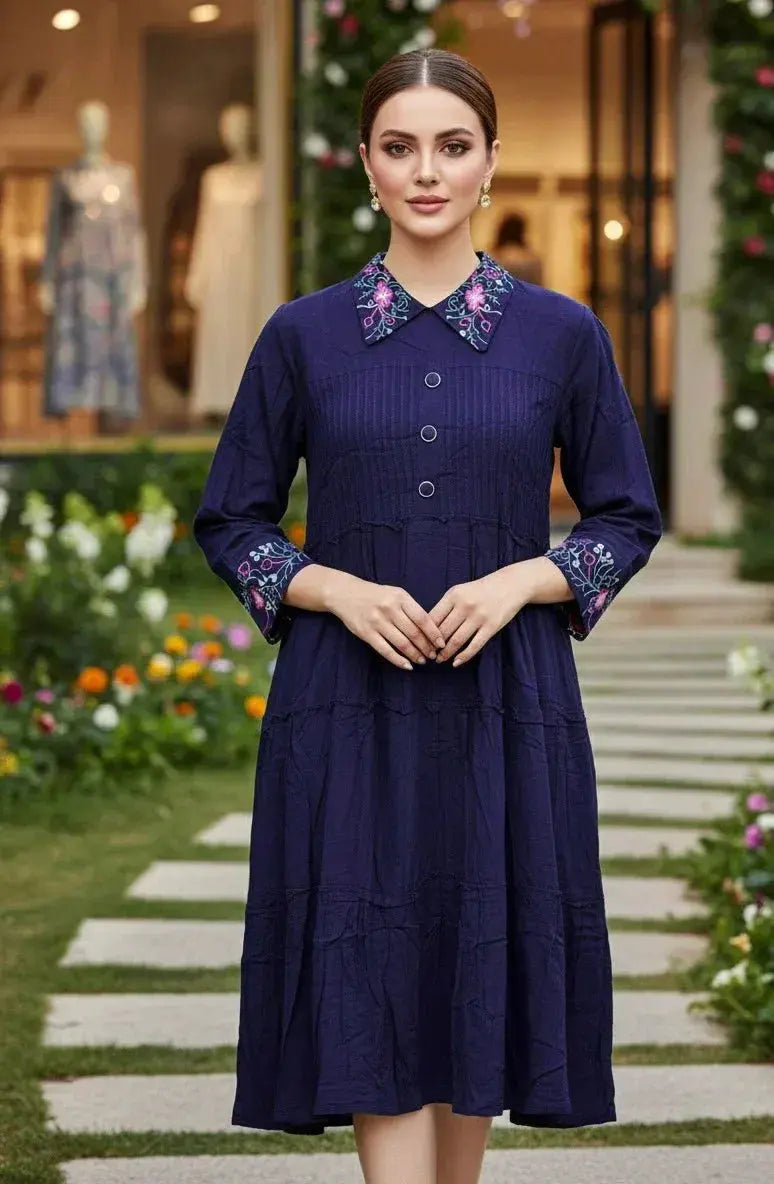 Woman wearing a navy blue dress with floral embroidered collar and cuffs, standing in a garden.