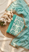 A folded embroidered fabric piece with a turquoise and white floral pattern, styled with a sheer turquoise dupatta and white flowers on a woven mat over a cream fabric background.